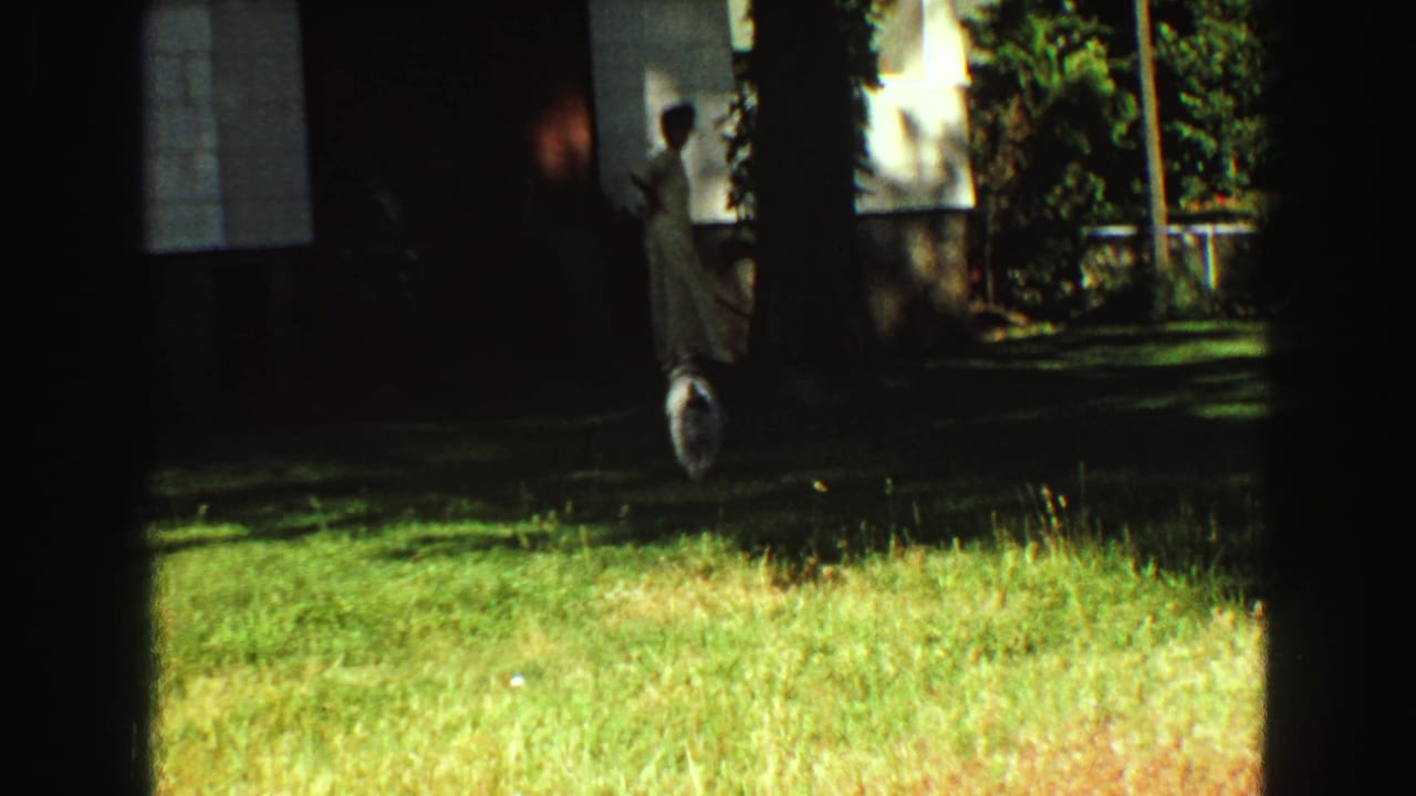 1968 Vancouver home-movie frame with a woman in a light dress near a house and a small white dog on the lawn.