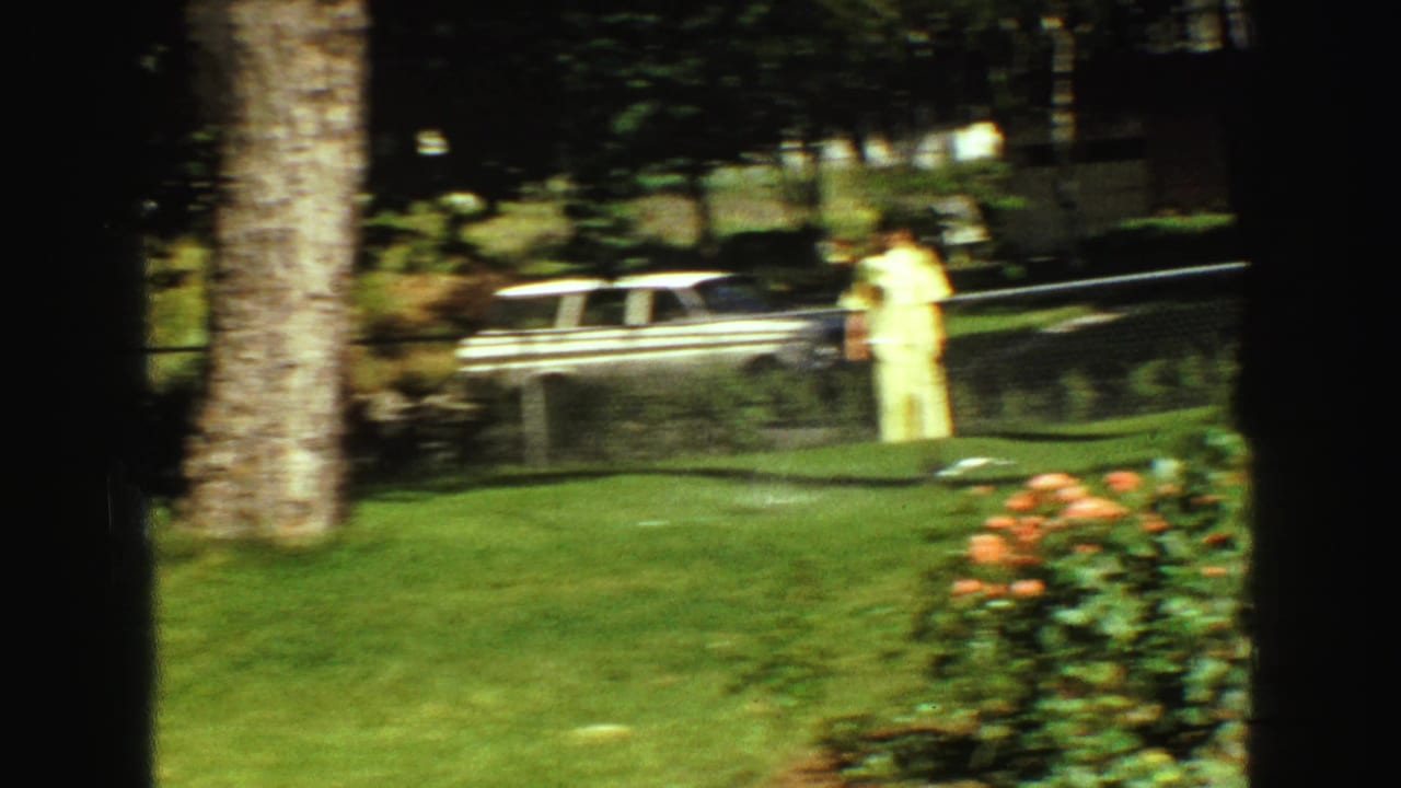Soft 8mm frame from Vancouver in 1968 showing a woman in a pale dress by the garden, with rose bushes, fence, and station wagon behind her.