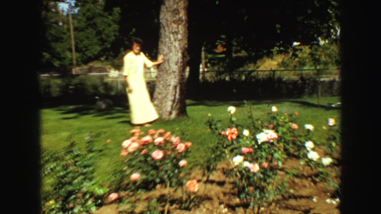 Woman in a pale yellow dress stands by a tree near rose bushes in a sunny yard.