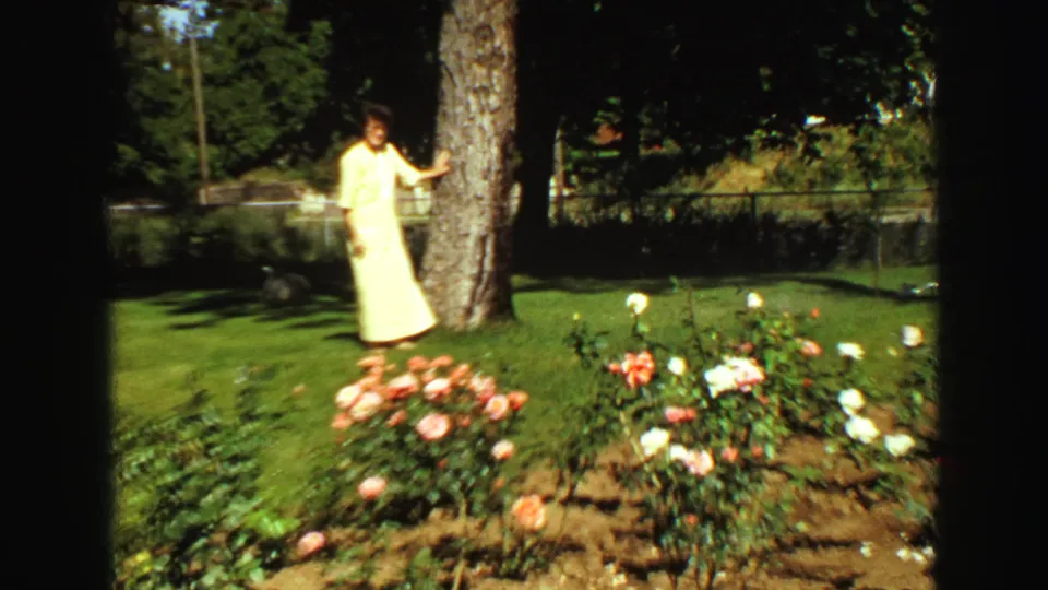 In 1968 Vancouver home-movie footage, a woman in a pale yellow dress stands beside a tree near rose bushes on a lawn.