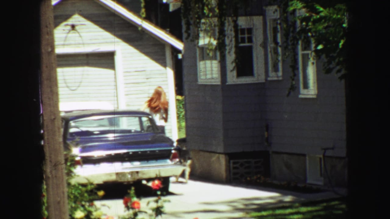 Woman near the rear of a blue sedan by a gray house and white garage.