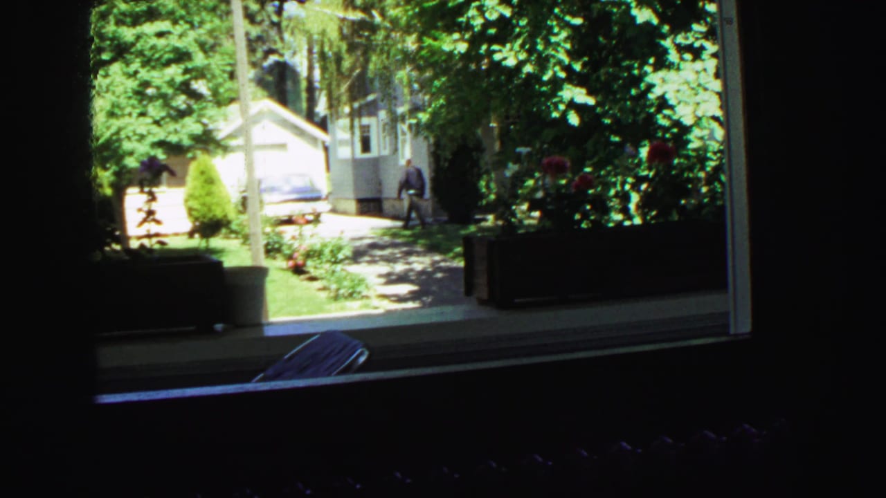 Wide 1968 Vancouver home-movie view from inside or behind a window, looking toward a residential driveway with a parked car and distant figure.