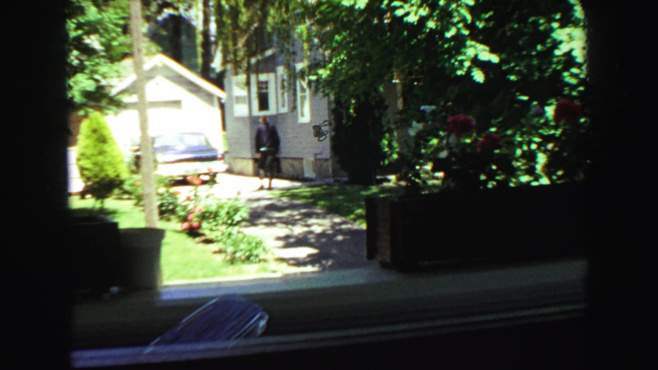 1968 Vancouver residential scene seen through a dark window frame, with a driveway, blue car, gray house, red flowers, and distant figure.