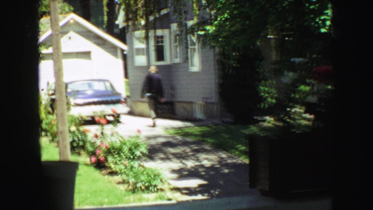 Man walking up a driveway toward a gray house and parked blue sedan.