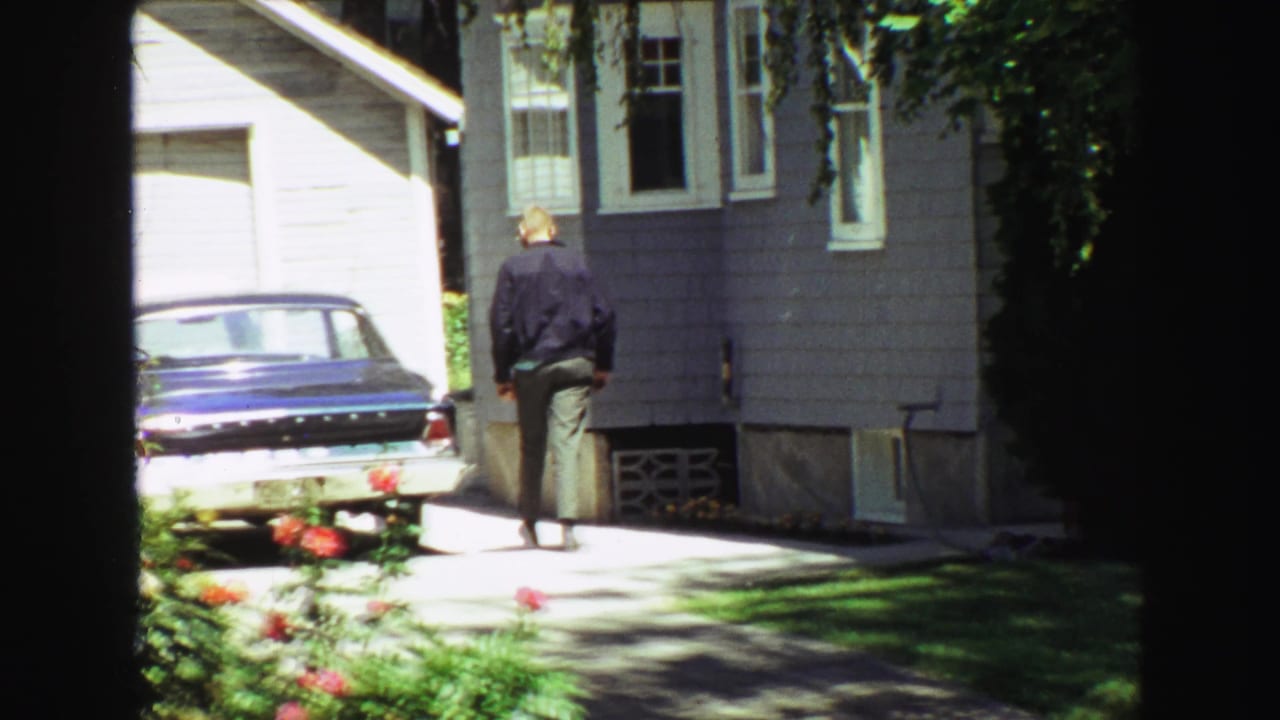 1968 Vancouver backyard view with a man approaching a gray-sided house, a blue sedan in the driveway, and garden flowers in foreground.