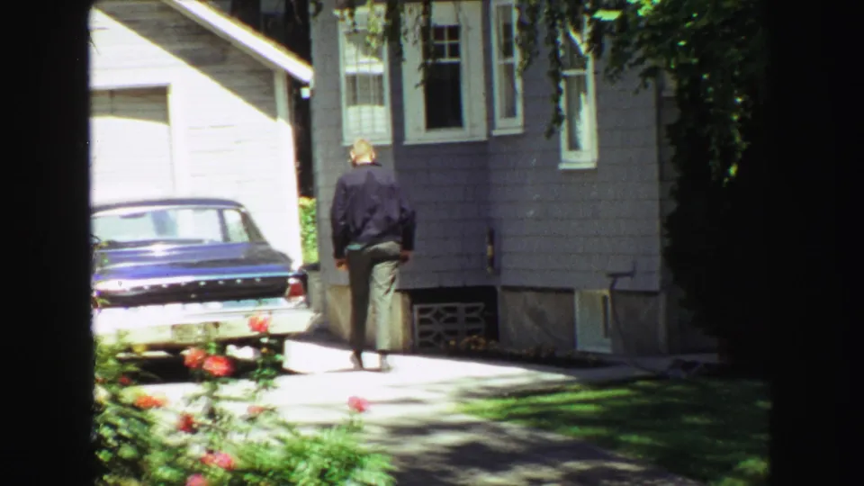 1968 Vancouver backyard view with a man approaching a gray-sided house, a blue sedan in the driveway, and garden flowers in foreground.