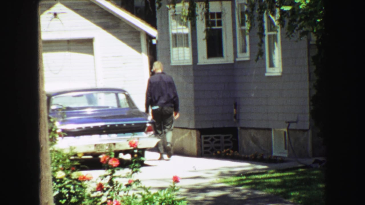 Home-movie frame from 1968 Vancouver showing a man beside a driveway sedan, with gray house siding, garage door, flowers, and deep window framing.