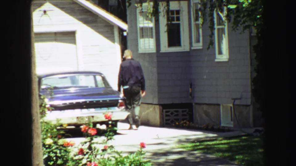 Home-movie frame from 1968 Vancouver showing a man beside a driveway sedan, with gray house siding, garage door, flowers, and deep window framing.