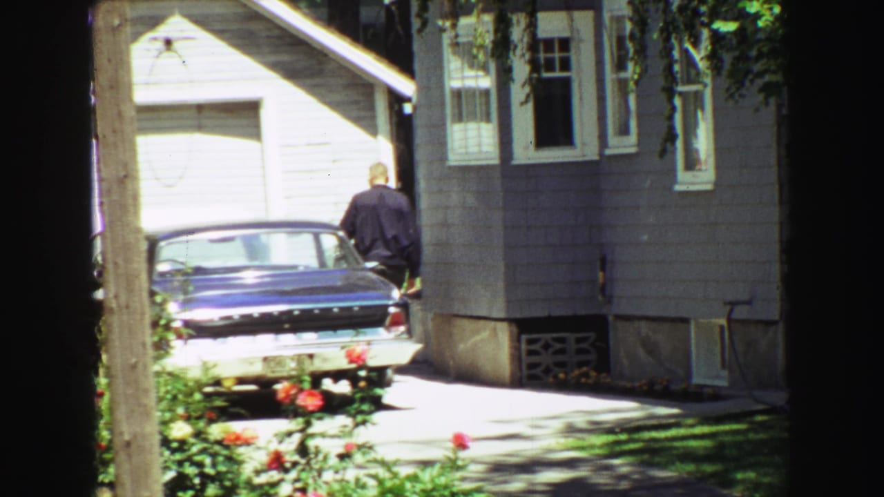 Final 1968 Vancouver frame with a man near a parked blue sedan, gray residential siding, white garage door, roses, and shaded lawn.