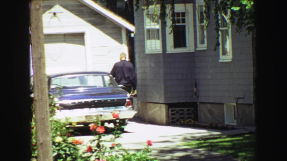 Final 1968 Vancouver frame with a man near a parked blue sedan, gray residential siding, white garage door, roses, and shaded lawn.