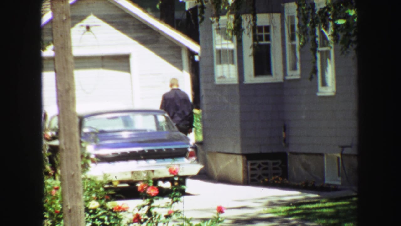 In supplied 1968 Vancouver home-movie footage, a man in a dark jacket walks toward a blue sedan parked beside a gray-sided house with roses and tree shade in the foreground.