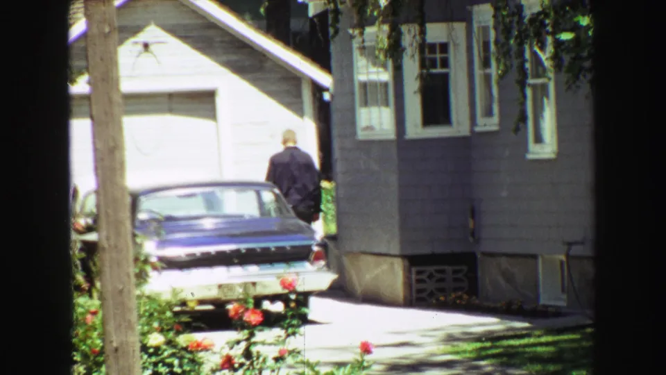 In supplied 1968 Vancouver home-movie footage, a man in a dark jacket walks toward a blue sedan parked beside a gray-sided house with roses and tree shade in the foreground.