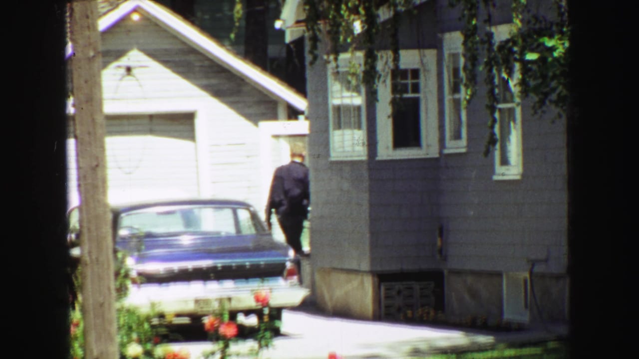 Man near the rear of a blue sedan beside a sunlit house.