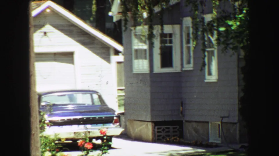 The driveway and parked blue sedan remain in view beside the gray-sided residential exterior, framed by a dark post and bright garden foliage.