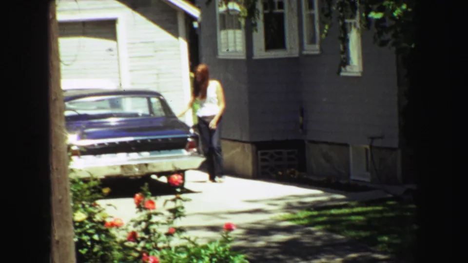 A woman in a white top and jeans appears near the parked blue sedan beside the gray house, continuing the supplied 1968 Vancouver home-movie driveway scene.