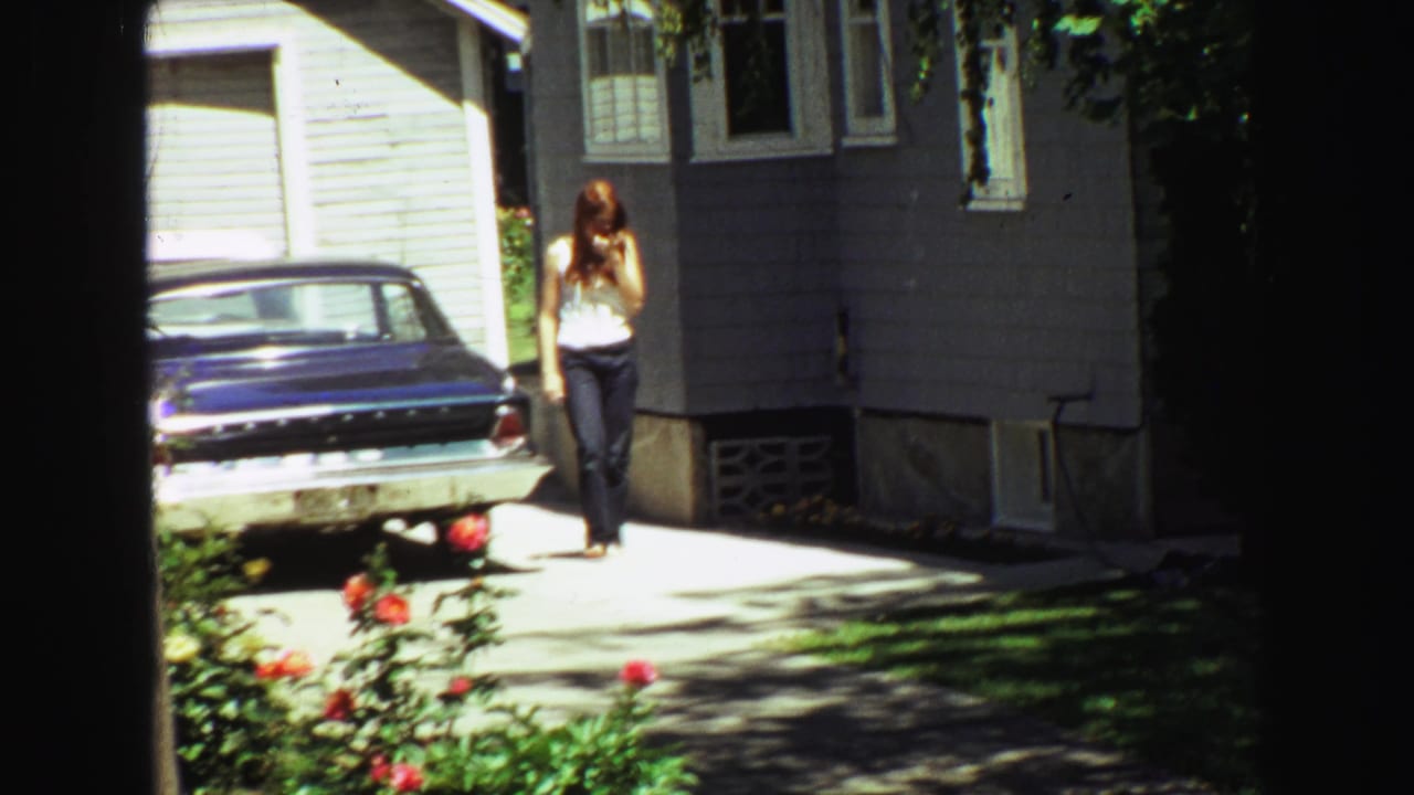 Woman walking across the driveway beside a parked blue car and house exterior.