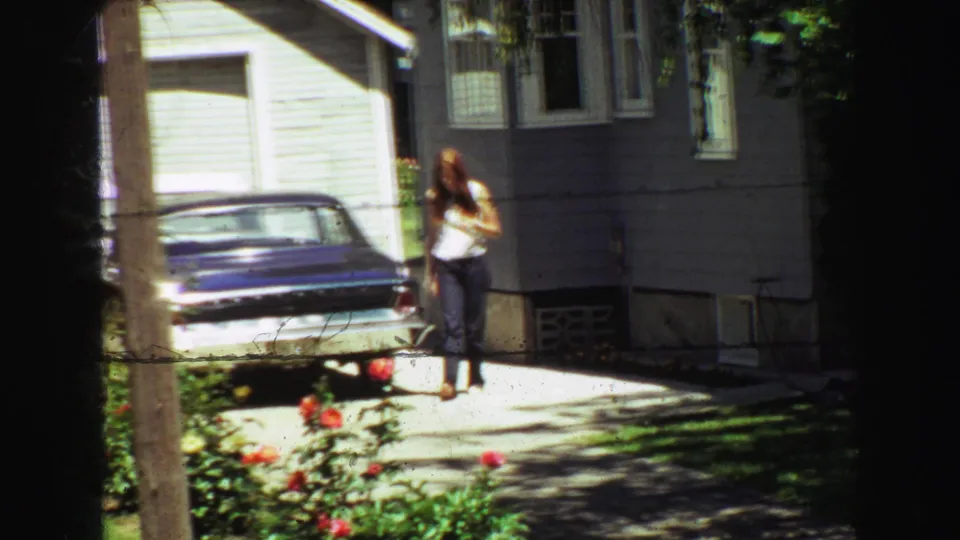Home-movie frame showing a young woman near a blue sedan and residential driveway, with red roses and a wooden post in the foreground.