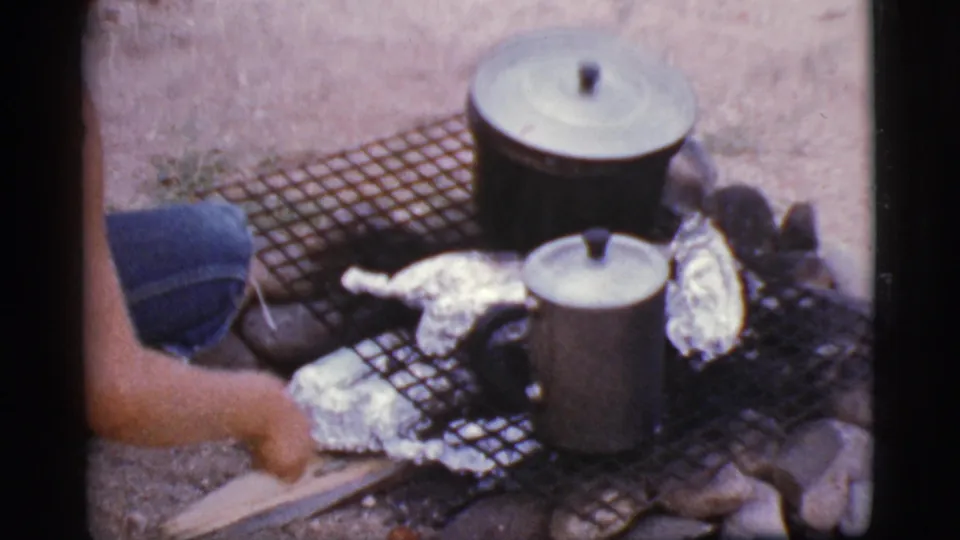 The home-movie frame shows a crouched person adjusting items near foil packets, pots, and a grill over a small outdoor fire.