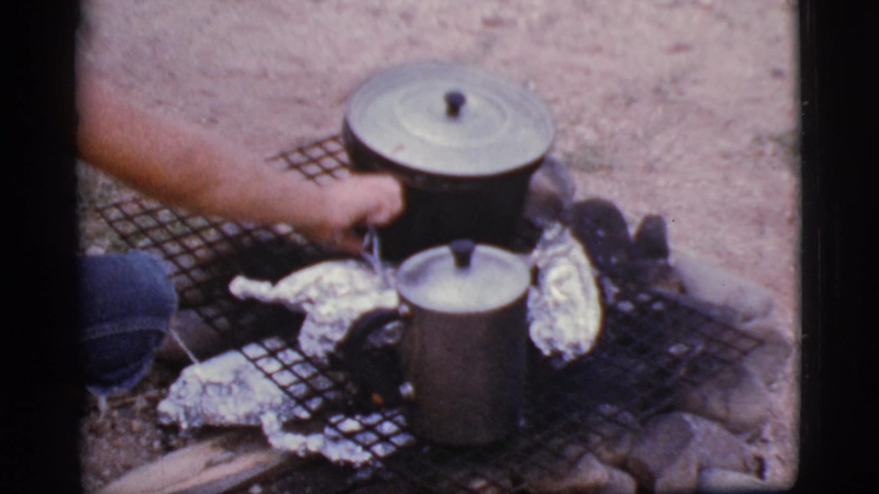 Hand near lidded cookware and foil-wrapped food on a wire grill over a small campfire.