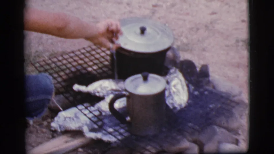 A hand uses a utensil around lidded cookware, foil packets, and smoke on a campfire grill in Cottonwood, Arizona, 1968.