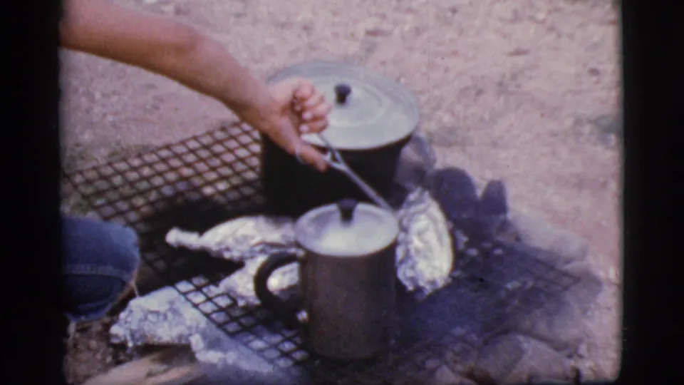 A close 8mm view shows a hand checking foil-wrapped food beside lidded cookware on a wire grate in 1968 Cottonwood.