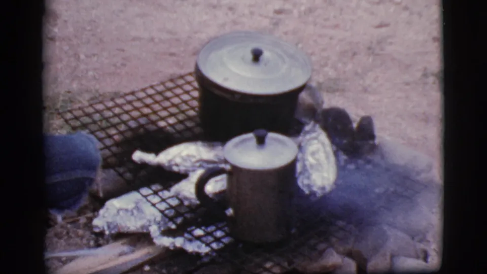 The campfire cooking arrangement remains visible with lidded pots, a metal mug, foil-wrapped food, smoke, and sandy ground.