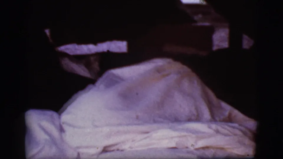 1968 Cottonwood, Arizona 8mm frame showing rumpled white covers with a raised shape and deep shadows around the bed.