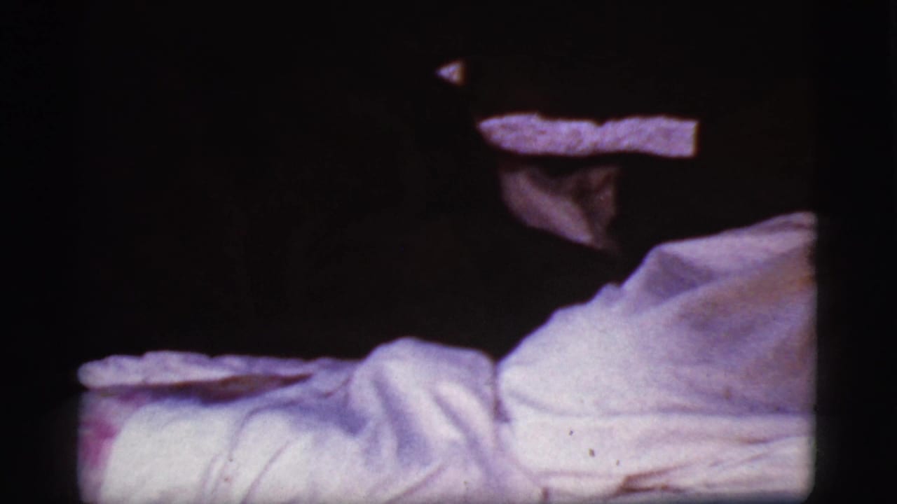 Dim cabin interior with a figure lying under a white blanket beside a dark wall.
