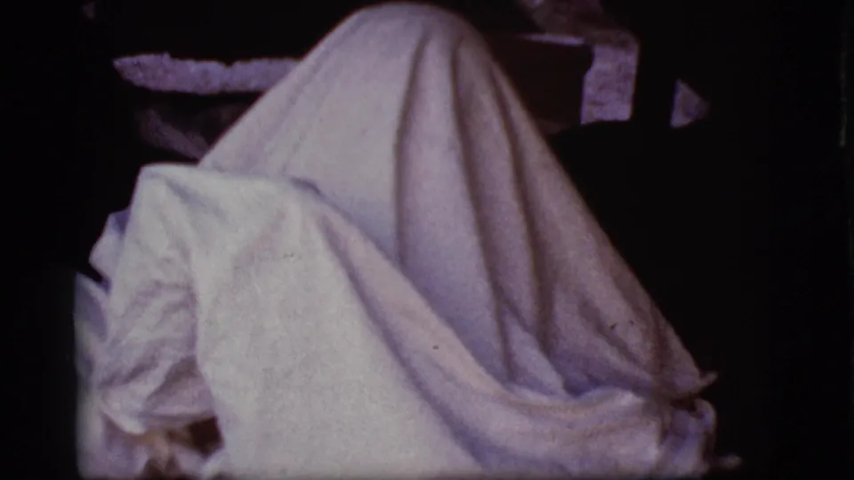 A sheet-covered figure bends forward in a dim Cottonwood, Arizona home-movie interior from 1968.