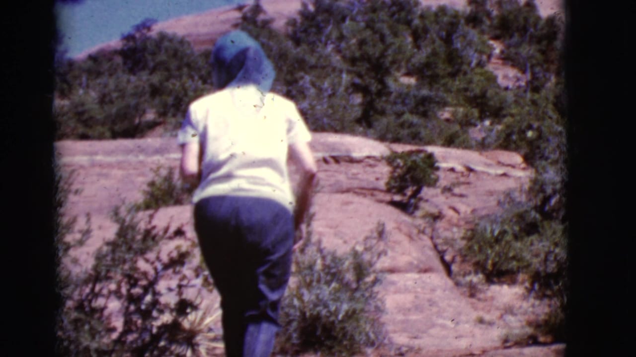 Woman in a blue head scarf walks uphill over red rock in desert brush.