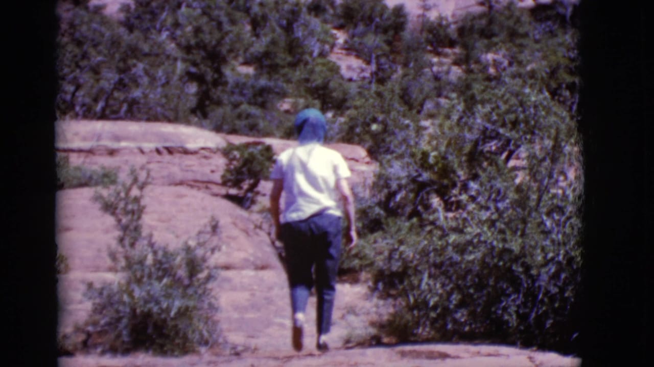 In Arizona 1968 home-movie footage, a person in a light shirt and blue head covering walks away on sunlit rock among desert shrubs.
