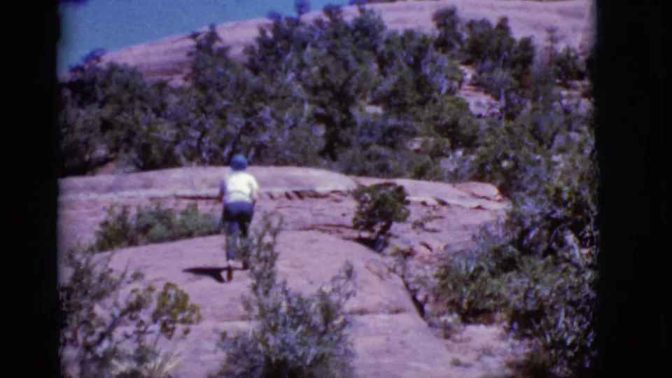 The hiker appears smaller in frame while moving uphill across a rocky Arizona landscape under bright sky in 1968 home-movie footage.