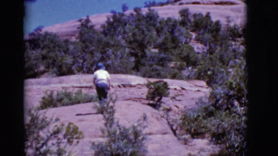 In 1968 Arizona home-movie footage, a woman in a white shirt walks uphill across smooth red rock and scrub.