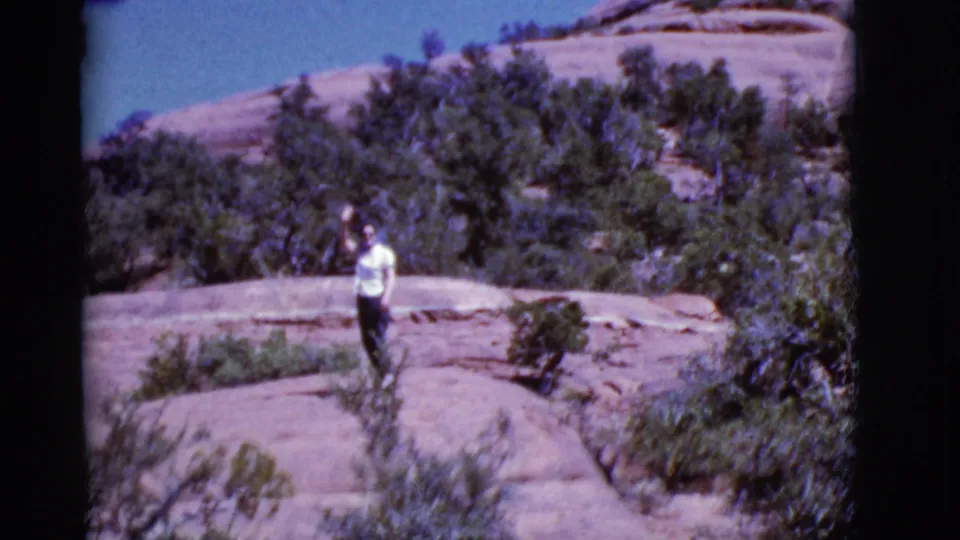 A woman in a white shirt faces the camera on a sunlit Arizona rock ledge, surrounded by desert vegetation.
