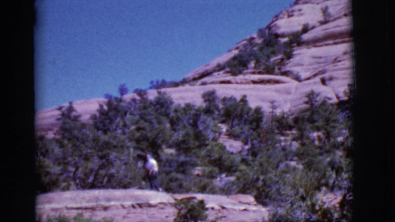 Man in a white shirt walks on a rocky desert ledge below sandstone slopes.