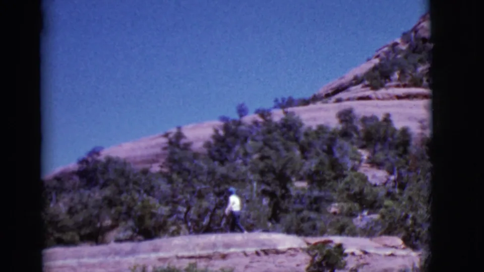 The 1968 Arizona scene shows a man in a white shirt moving along a sunlit rocky path against scrub and sandstone.