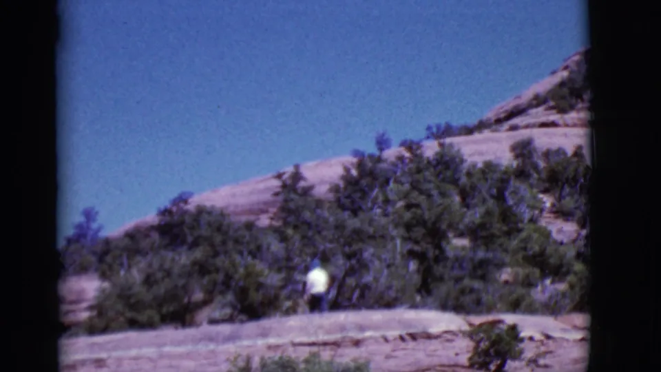 A white-shirted man is partly framed by desert vegetation while moving along a rocky Arizona trail in 1968 footage.