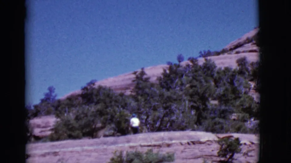 In documented 1968 Arizona footage, a distant person in white walks across a sunlit rocky slope bordered by trees and brush.