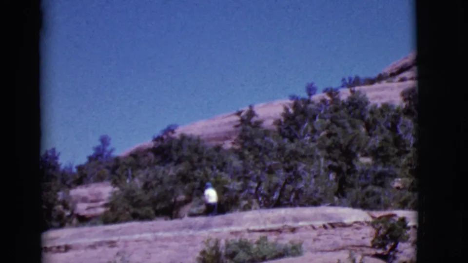 A distant walker in a white shirt and hat crosses a pale rock ledge in the Arizona desert landscape.