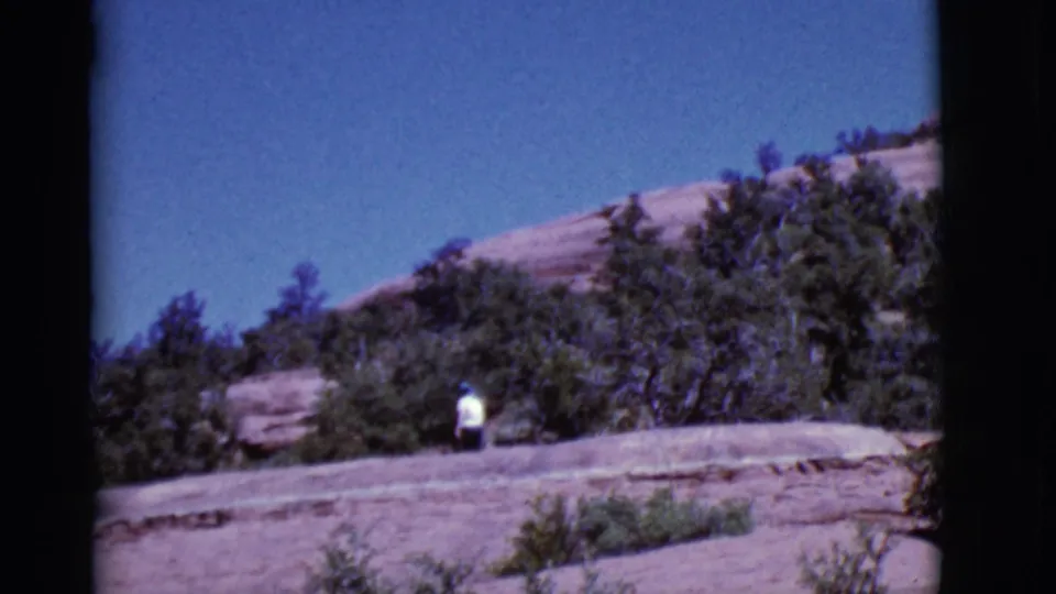 The 8mm frame shows a small figure in white moving below trees and exposed rock in Arizona.