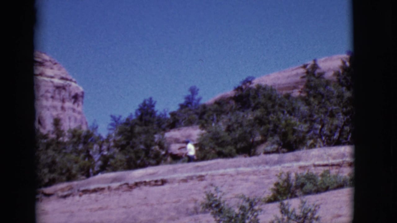 Person in a white shirt walking below red rock cliffs and green trees.