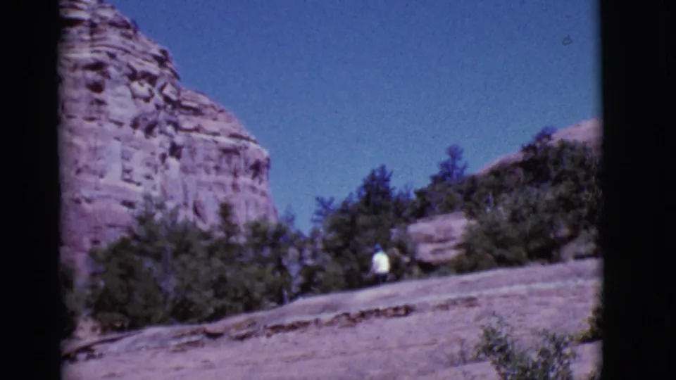 Arizona home-movie frame from 1968 with a small figure walking near shrubs below a red cliff face.