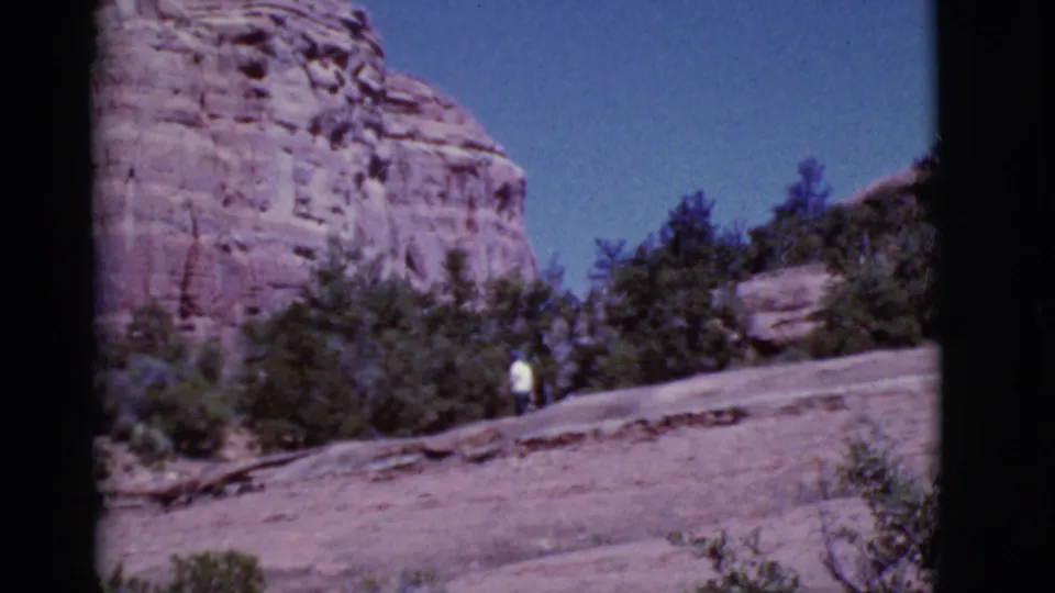 Arizona 1968 home-movie frame of a lone walker in a white shirt crossing a rocky trail under red cliffs.