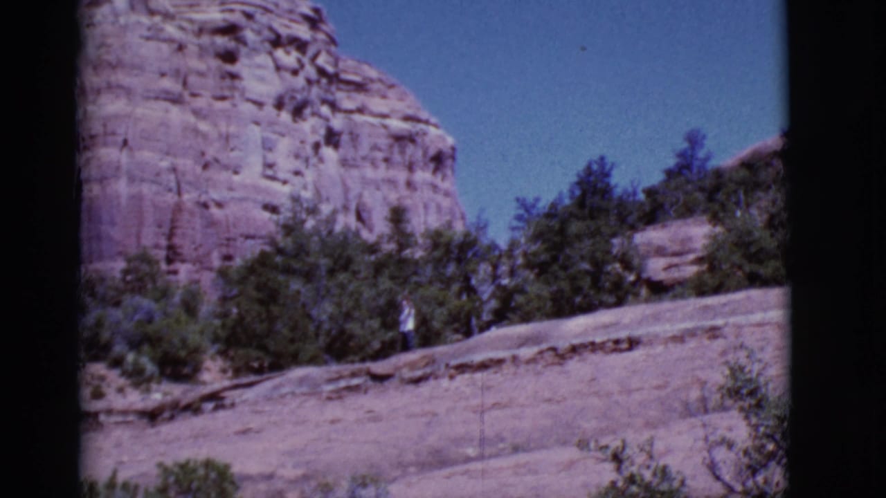Red rock desert slope with shrubs and a cliff under blue sky.