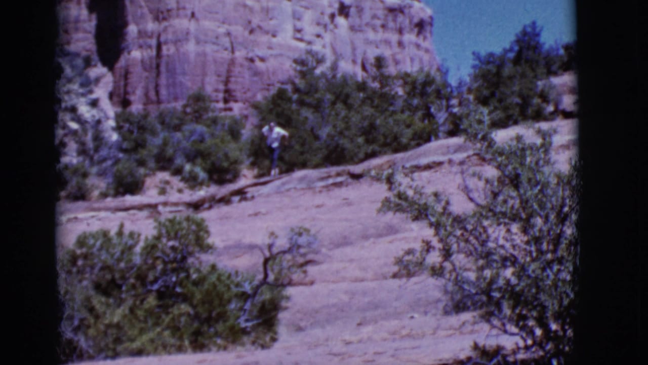 Person walking uphill on a rocky desert slope with shrubs and red rock behind.
