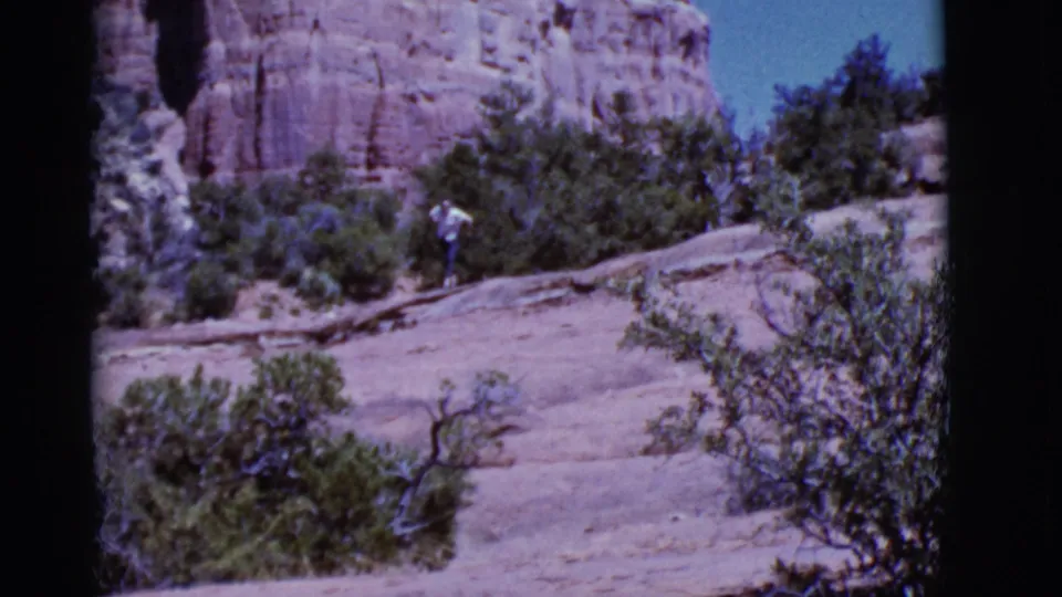 1968 Arizona 8mm frame showing a person in a light shirt hiking across a rocky desert slope with shrubs and red rock formations.