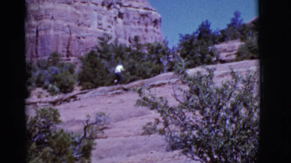 1968 Arizona home-movie frame showing the hiker moving through shrubs on a pale rock incline below red desert stone.