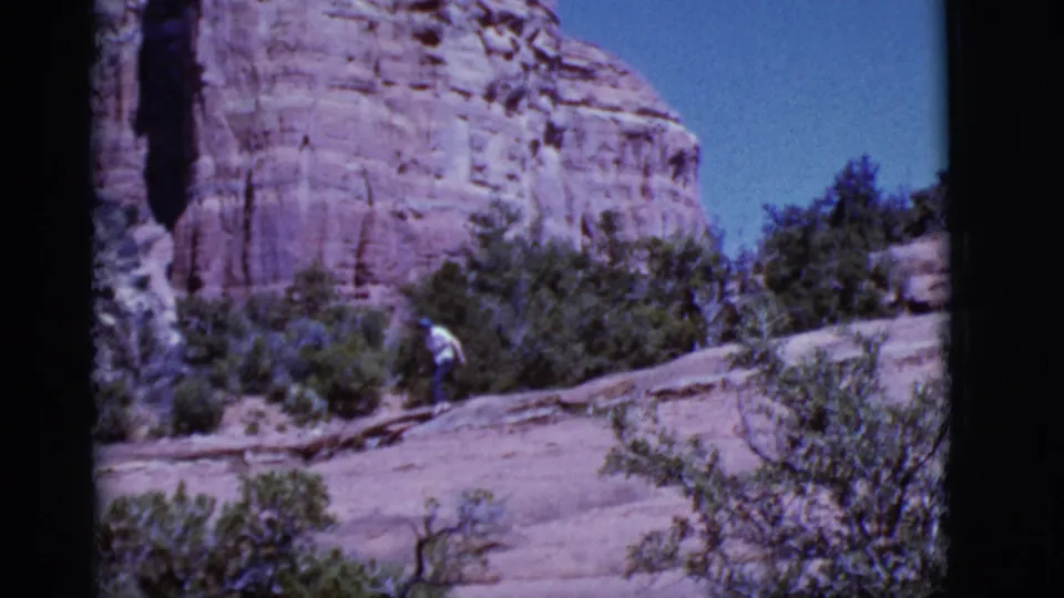 The hiker is seen on the Arizona slope near low shrubs, with red rock formations filling the background in vintage 8mm footage.