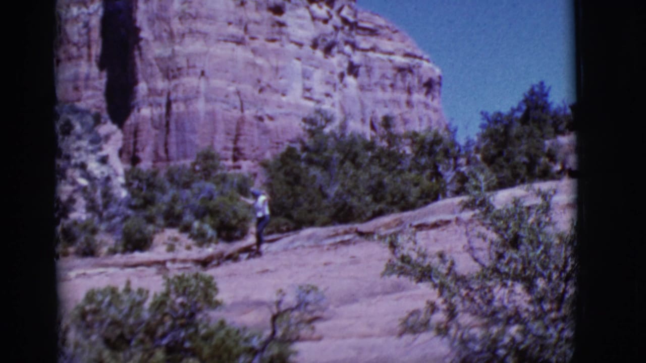 Woman walking on a rocky desert slope with red cliffs and shrubs behind her.