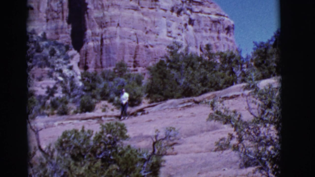 Woman walking near shrubs on pale rock with a red cliff in the background.